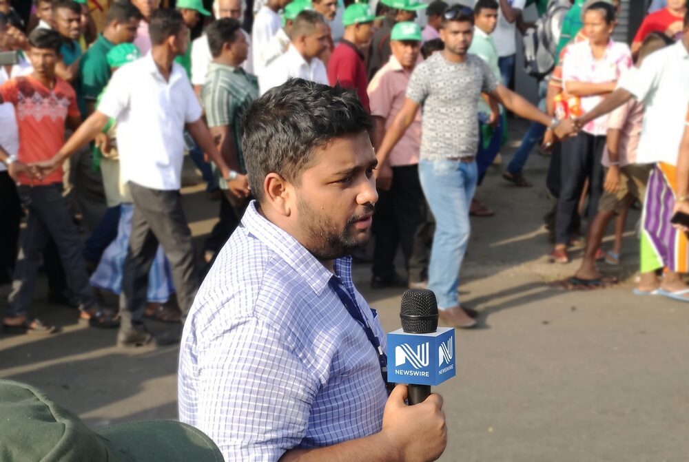 Azzam Ameen reporting from a street crowd, holding a microphone with a blue NewsWire mic flag